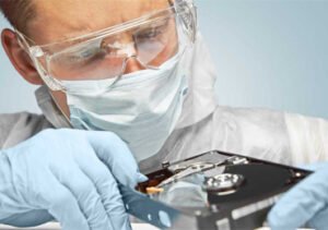 Technicien masqué avec lunettes et gants inspectant un disque dur.