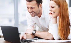 Deux personnes sourient devant un ordinateur portable dans un bureau.