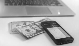 Smartphone with calculator showing 200 next to twenty-dollar bills on table, blurred laptop behind